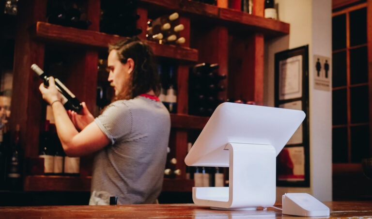 An electronic point of sale machine. In the background, a wine seller looks at a bottle of wine.